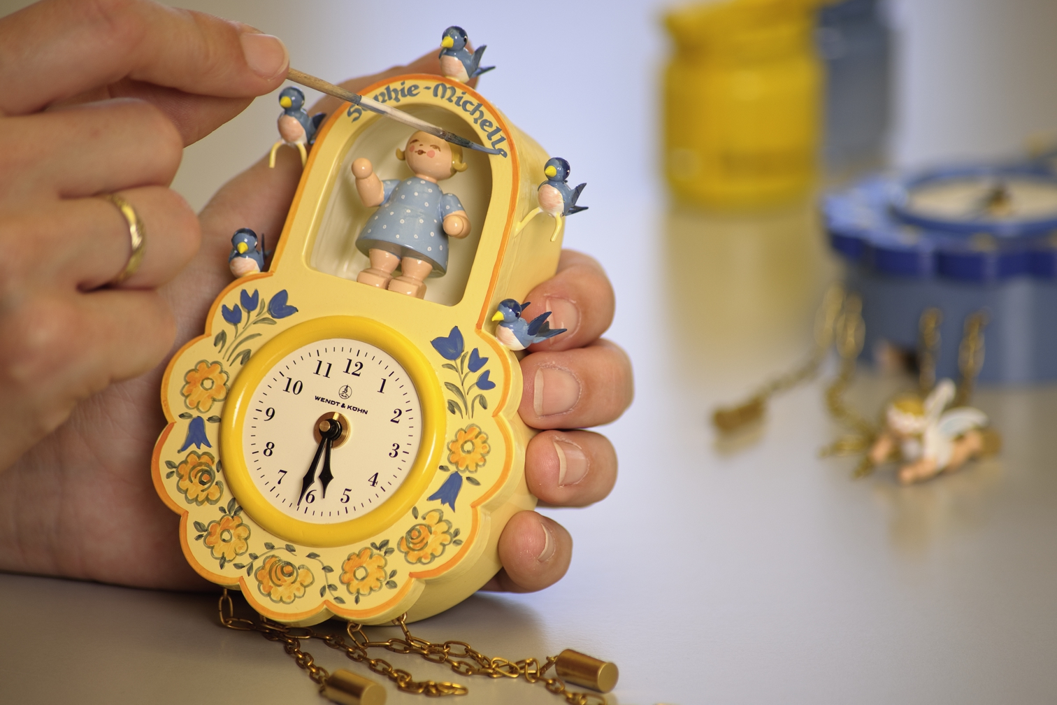 Nahaufnahme einer gelben Wanduhr von Wendt & Kühn in Blumenform mit einer Kinderfigur und blauen Vögeln, die von einer Person von Hand bemalt wird